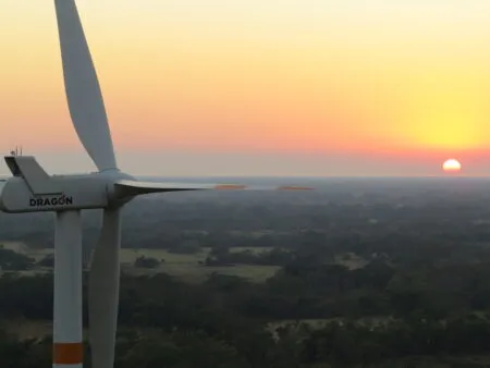 Wind turbine blade in the foreground with a colorful sunset and distant fields below.