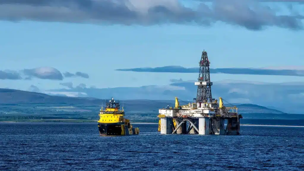 Offshore oil rig with a yellow tugboat nearby on calm blue water.
