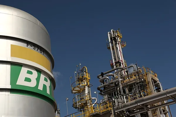 Large BR-branded storage tank beside an industrial refinery with yellow staircases and extensive piping against a clear blue sky.