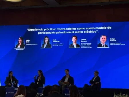 Panel discussion on a stage with four speakers seated in chairs, microphones in hand, against a blue geometric backdrop with a large title screen above.