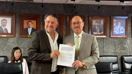Two men in suits smile while presenting a signed document in a formal office setting with portraits on the stone wall behind them.