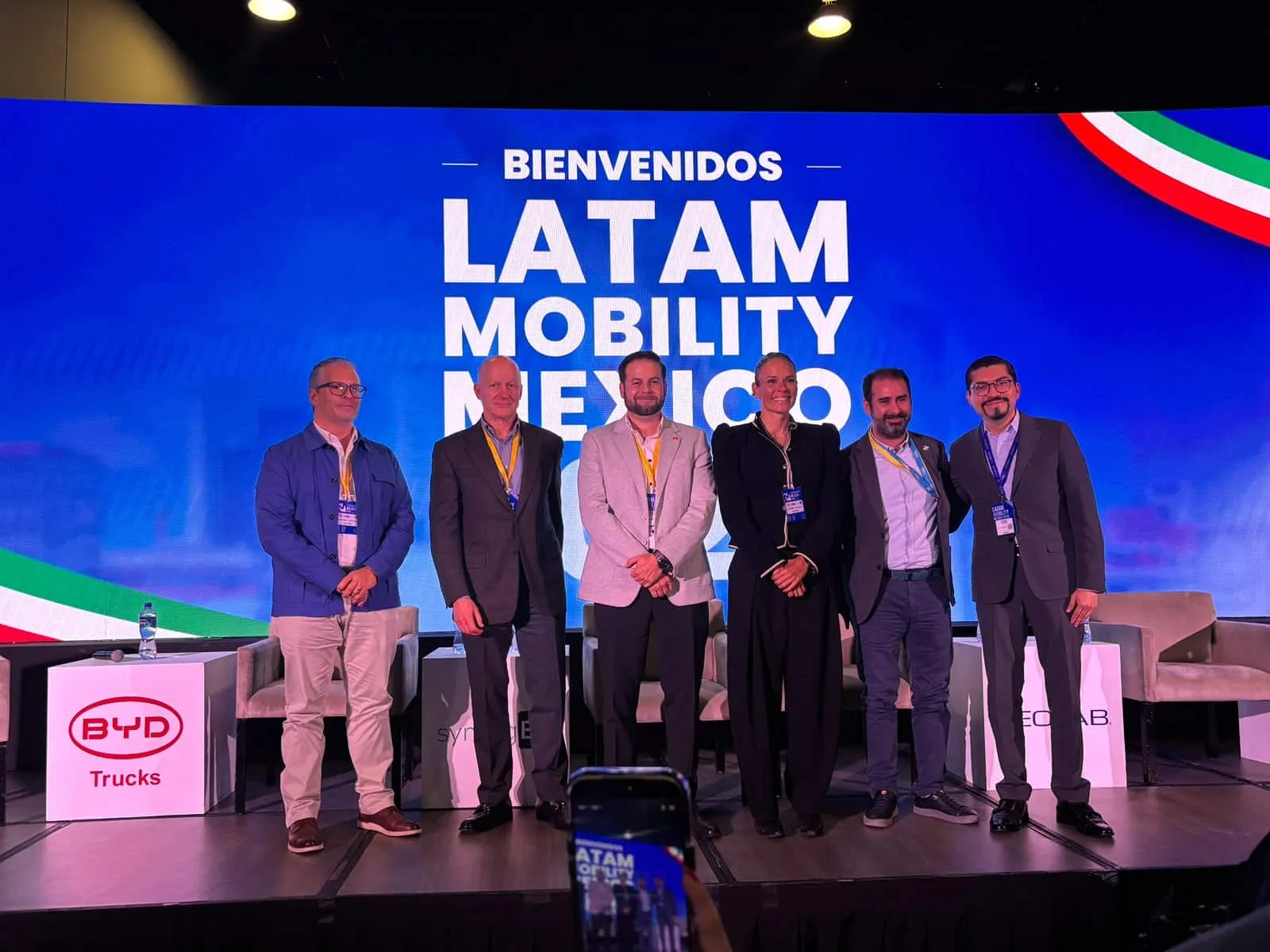 Six professionals pose on a stage in front of a bright blue LATAM MOBILITY MEXICO backdrop, badges visible.
