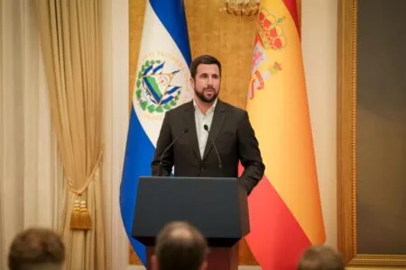 Man in a suit giving a speech at a podium, flanked by El Salvador and Spain flags.