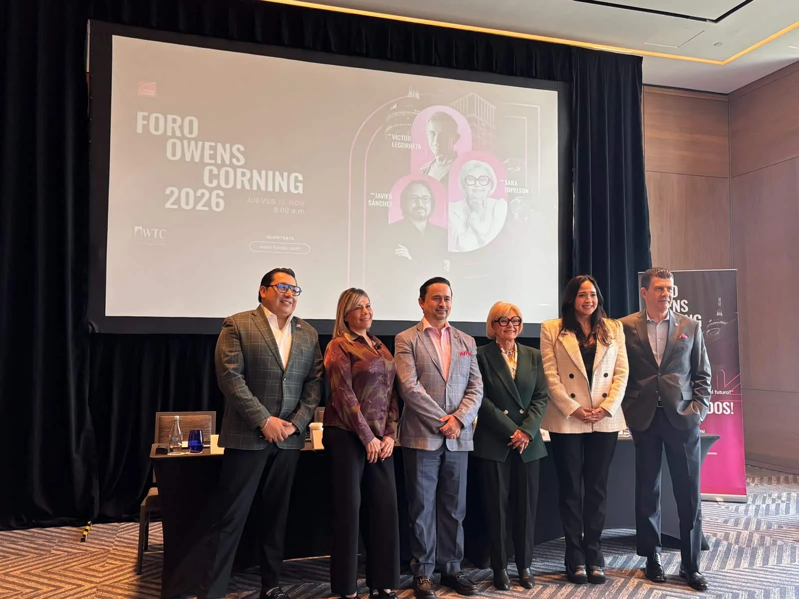 Six professionals in business attire pose on a conference stage in front of a large screen displaying Foro Owens Corning 2026 poster and graphics behind them.