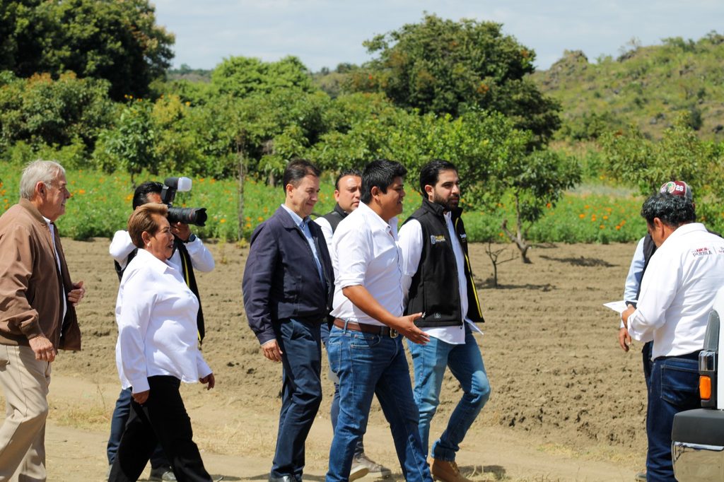 WhatsApp_Image_2026-10-27_at_114019_AM_1 - Petróleo y Energía Group of people walking in a dirt field with photographers and reporters following, outdoors among green plants and trees.
