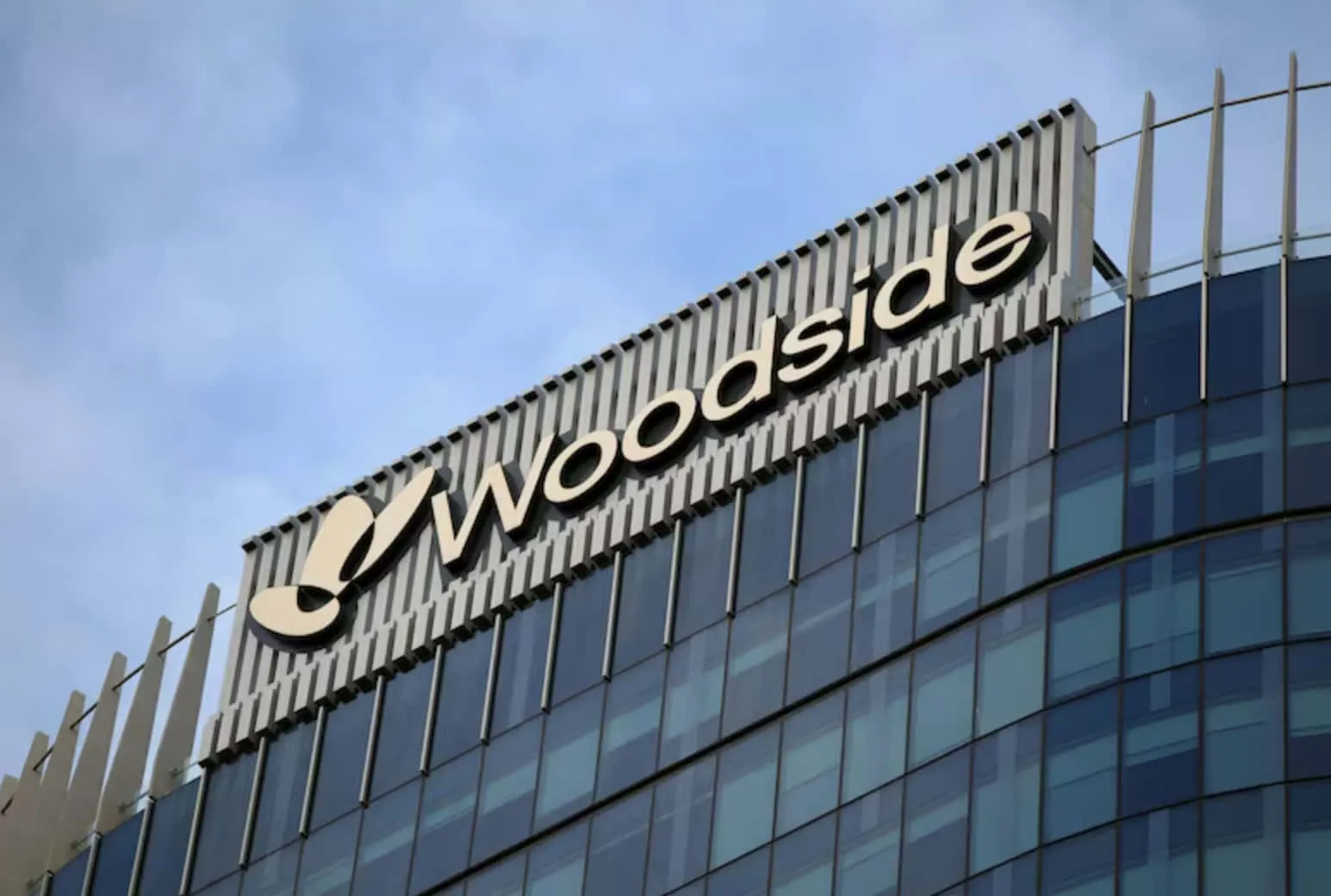 Woodside company sign mounted along the top of a glass-office building against a blue sky.