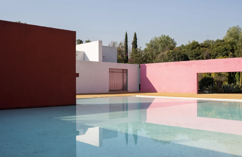 Colorful modern building with pink and red walls surrounding a reflective pool under a clear blue sky.