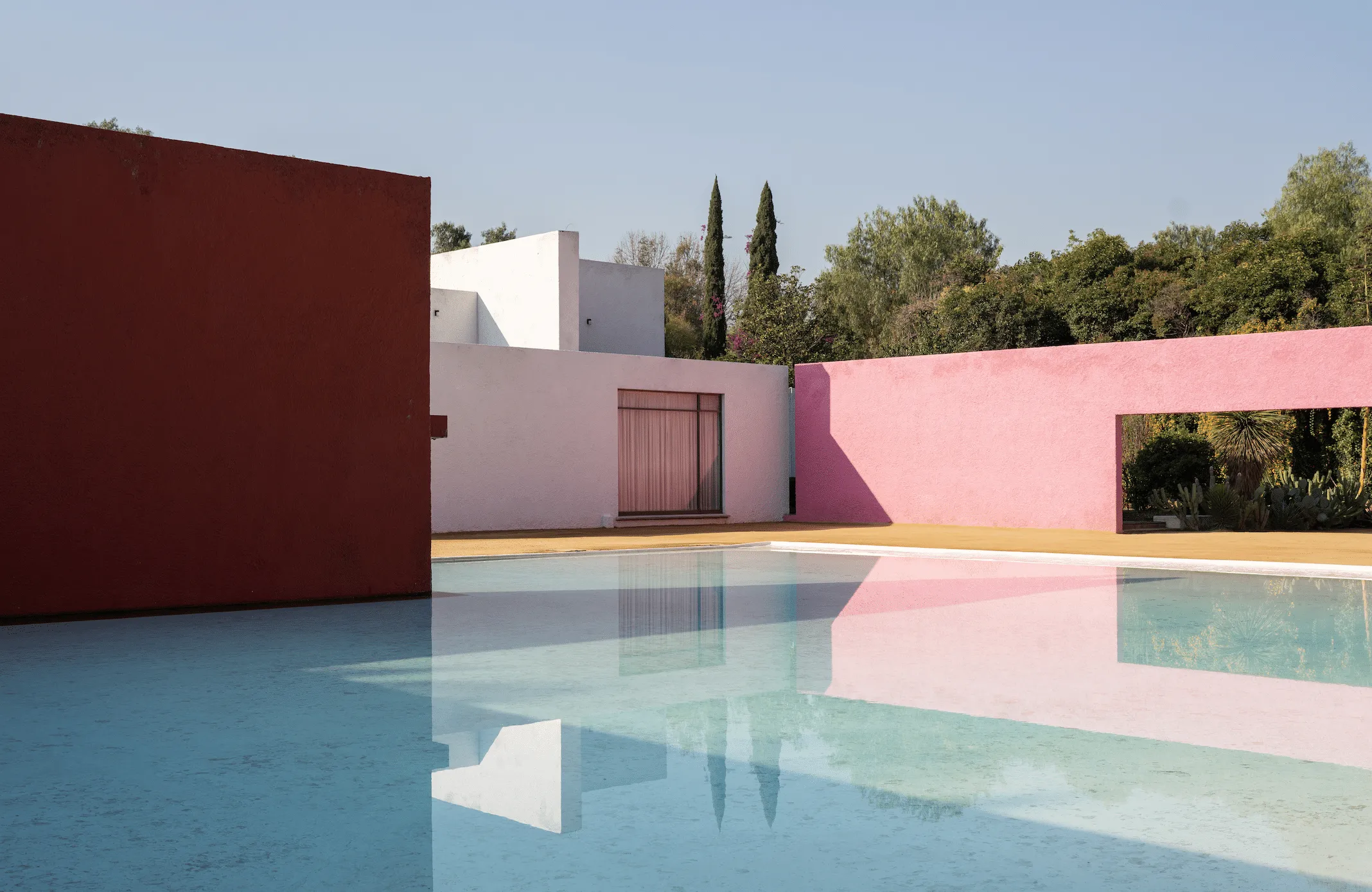 Colorful modern building with pink and red walls surrounding a reflective pool under a clear blue sky.