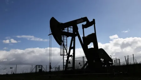Silhouette of an oil pumpjack in a fenced field against a blue, cloud-filled sky.