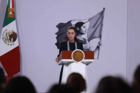 Bride at a podium: a formal speaker stands behind a wooden podium with a round Mexican seal, flanked by Mexican flags and a large black-and-white flag in the background, audience silhouettes in foreground.