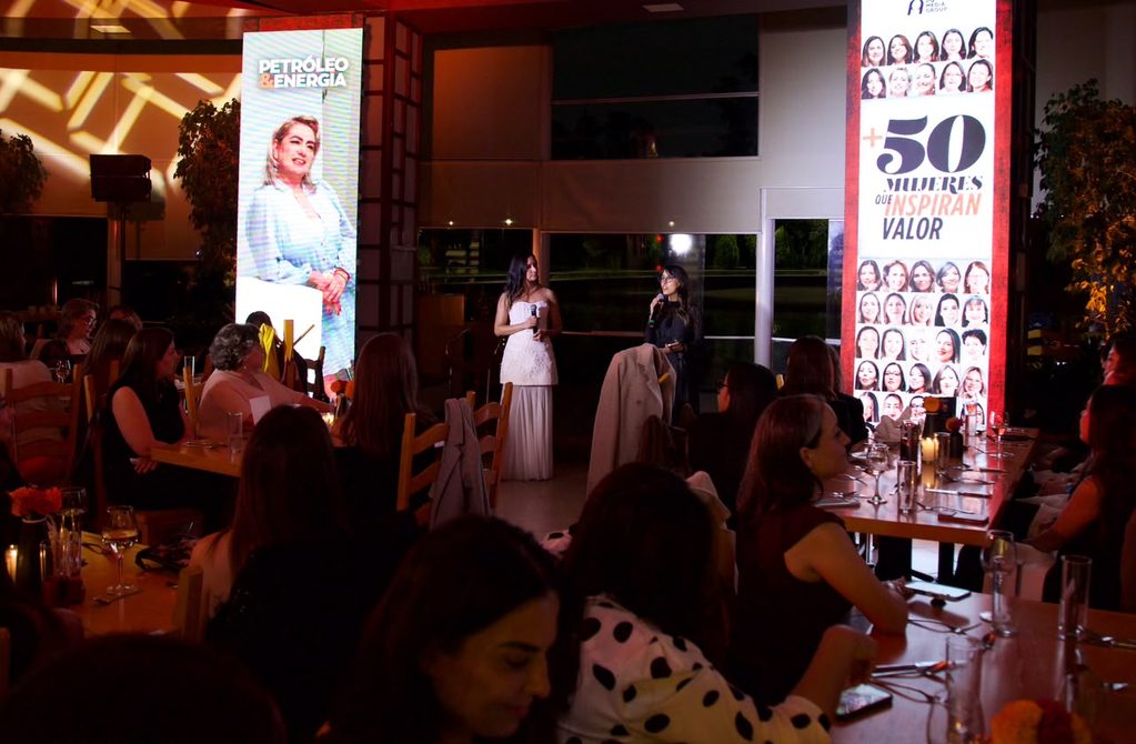 conductorasde50mujres - Petróleo y Energía Two women in evening dresses speak into microphones on a stage at a banquet, while guests sit at tables and tall banners frame the scene.