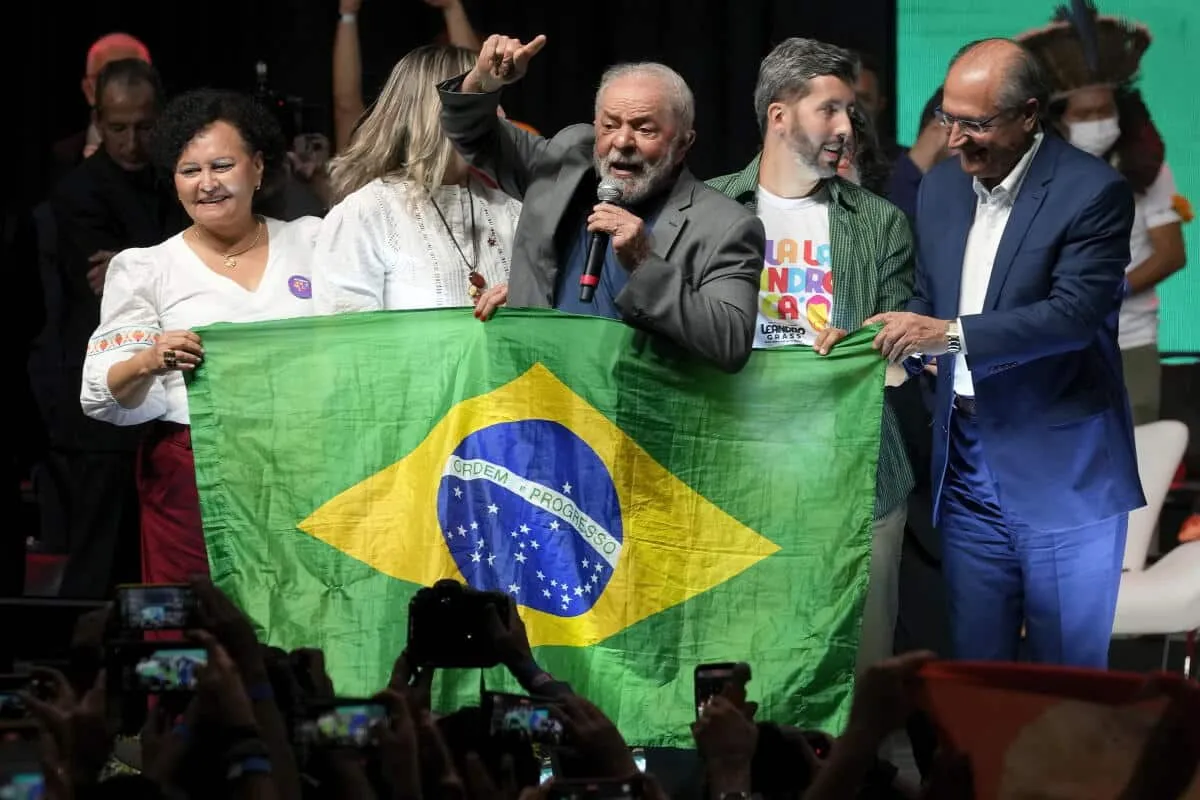 Group of people on stage holding a Brazil flag during a rally, with photographers in the crowd below.