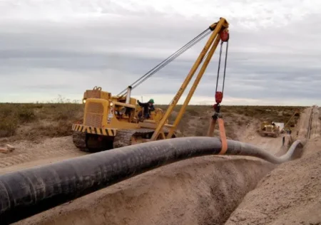 Heavy construction crane lifts a large black pipeline into a trench at a dirt site, with workers and equipment nearby