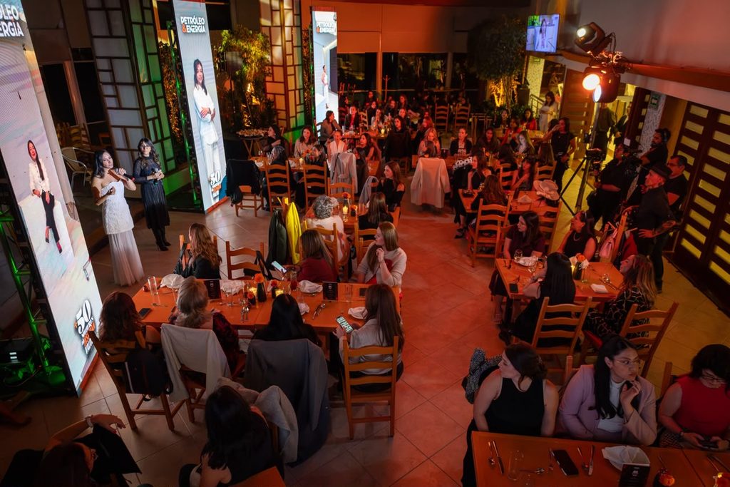 envetomujeres - Petróleo y Energía Audience at a formal dinner watches two women speak on a stage with banners in a warmly lit room at a banquet hall.
