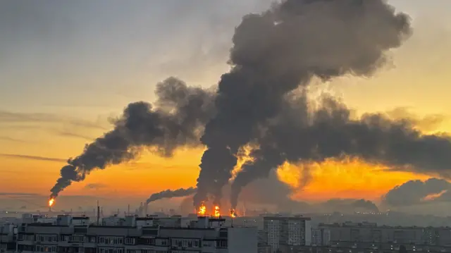 Industrial area at sunset with tall smokestacks emitting dark smoke and visible flames on the rooftops, creating an orange sky.