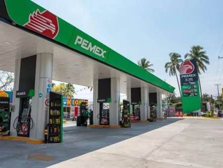Pemex gas station with a green canopy, multiple pump bays, and a price board under a clear blue sky with palm trees nearby.