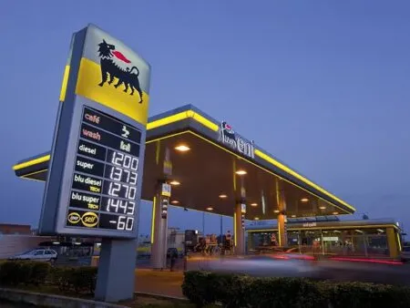 Gas station at dusk with a tall sign featuring a black lion logo; price board shows fuel types like diesel and super under a blue sky.