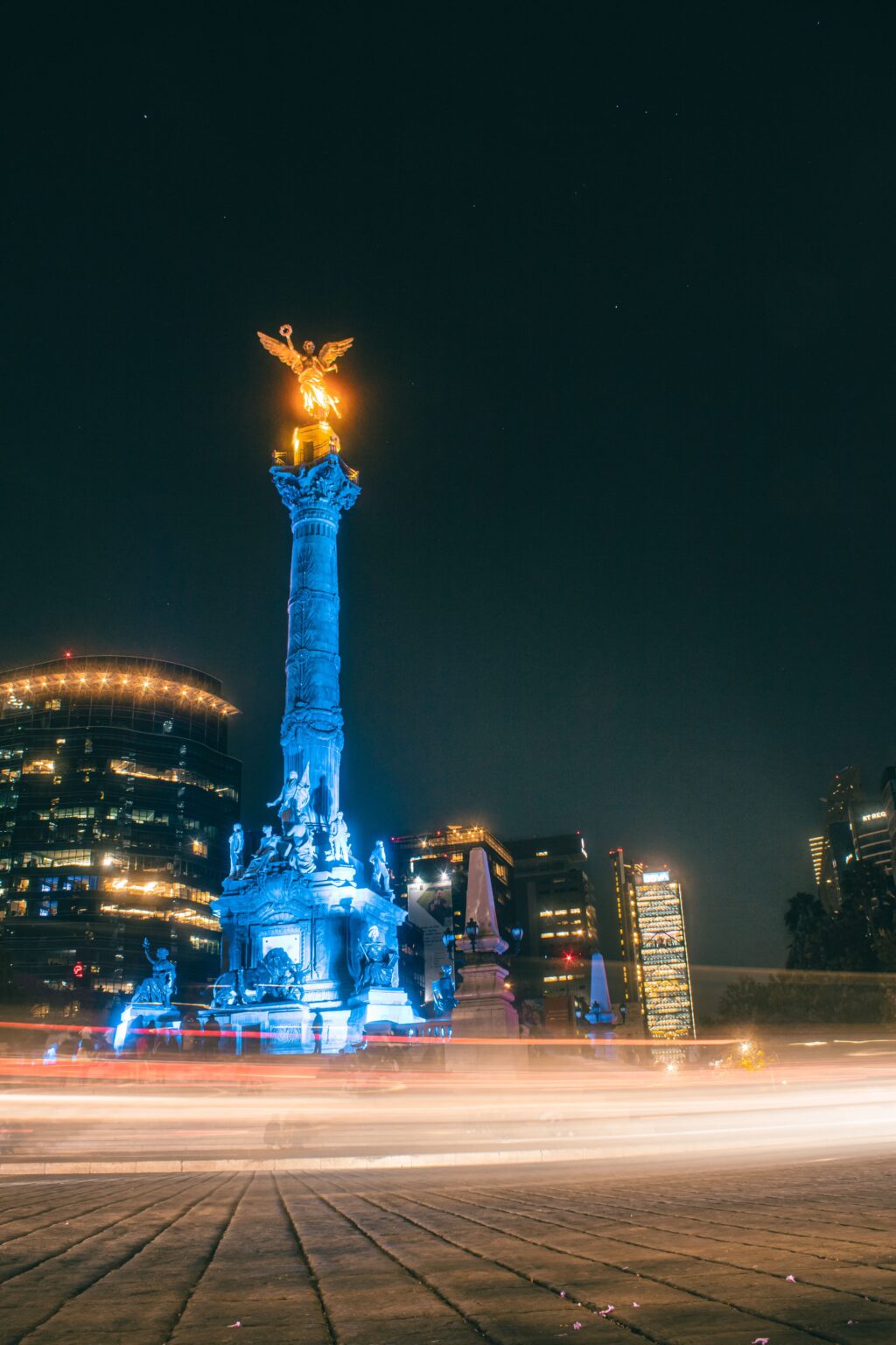 - Petróleo y Energía Nighttime view of a tall illuminated column with a golden winged statue on top, glowing blue with surrounding city lights in a plaza.