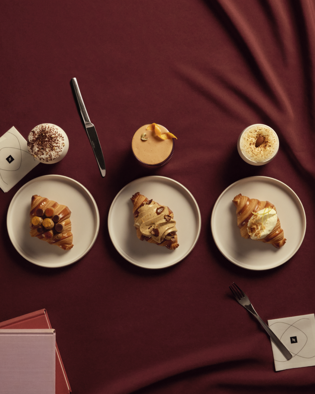 please distrub 2026-10-08 a las 11341 pm - Petróleo y Energía Top-down view of three pastries on white plates with a knife and drinks on a burgundy tablecloth.