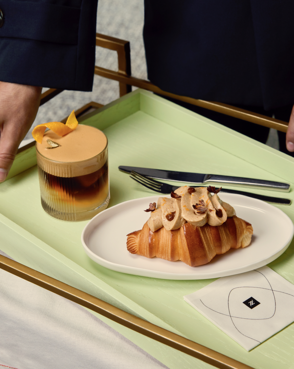 - Petróleo y Energía Croissant pastry topped with cream and chocolate garnishes on a white plate beside a layered iced coffee on a light green tray. A hand reaches toward the tray in a dark outfit behind the items.
