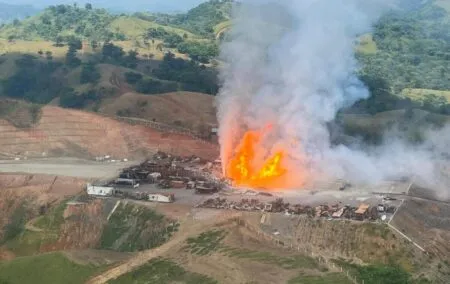 Industrial site on a hillside with a large explosion and orange flames shooting upward amid white smoke.