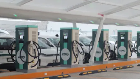Row of electric vehicle charging stations with hoses and cars parked at a charging bay area, labeled CARGADOR ELÉCTRICO (VEMO).
