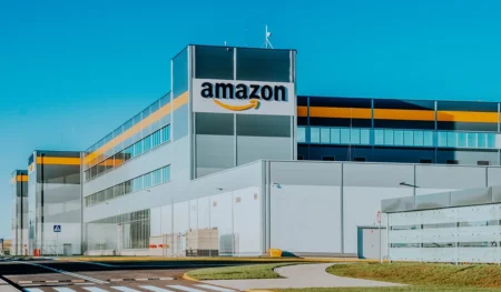 Amazon fulfillment center exterior with large logo, white building, orange accent stripe, and blue sky