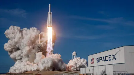 SpaceX rocket blasts off from launch pad with billowing smoke, flames, and a clear blue sky in the background, adjacent to a SpaceX hangar.