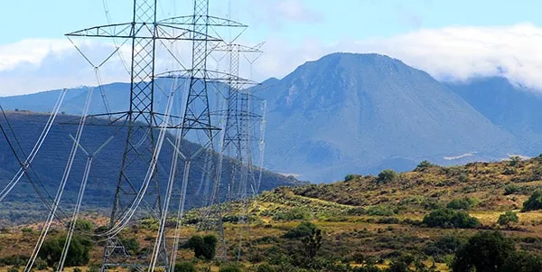 High-voltage transmission towers and lines stretch across a grassy hillside with a mountain range in the background.
