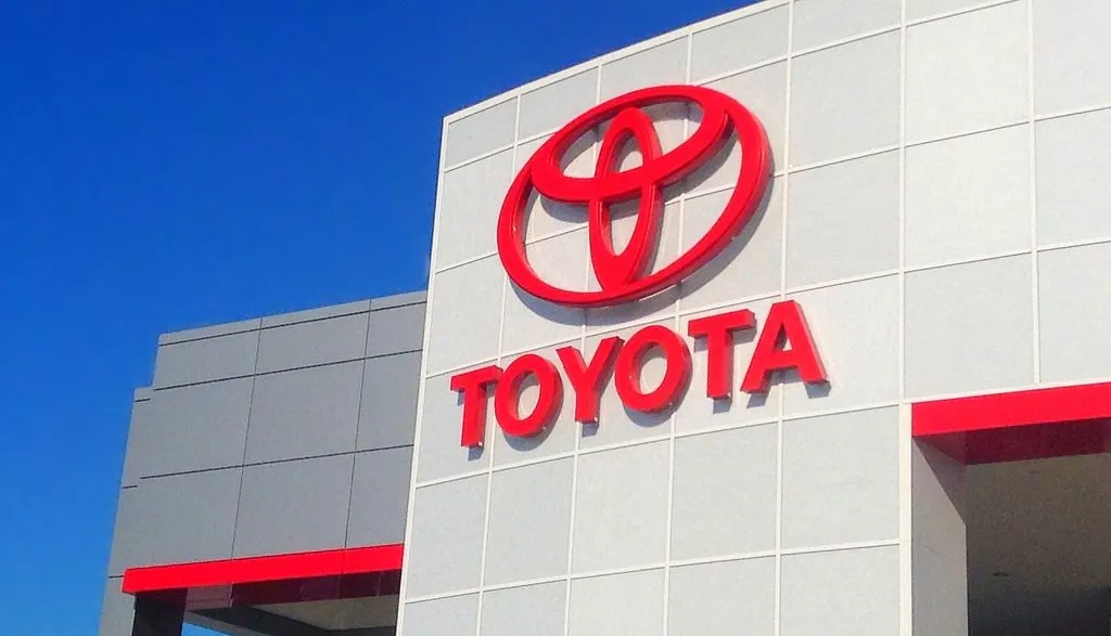 Toyota dealership exterior with large red Toyota emblem and sign on a light gray tiled facade against a blue sky